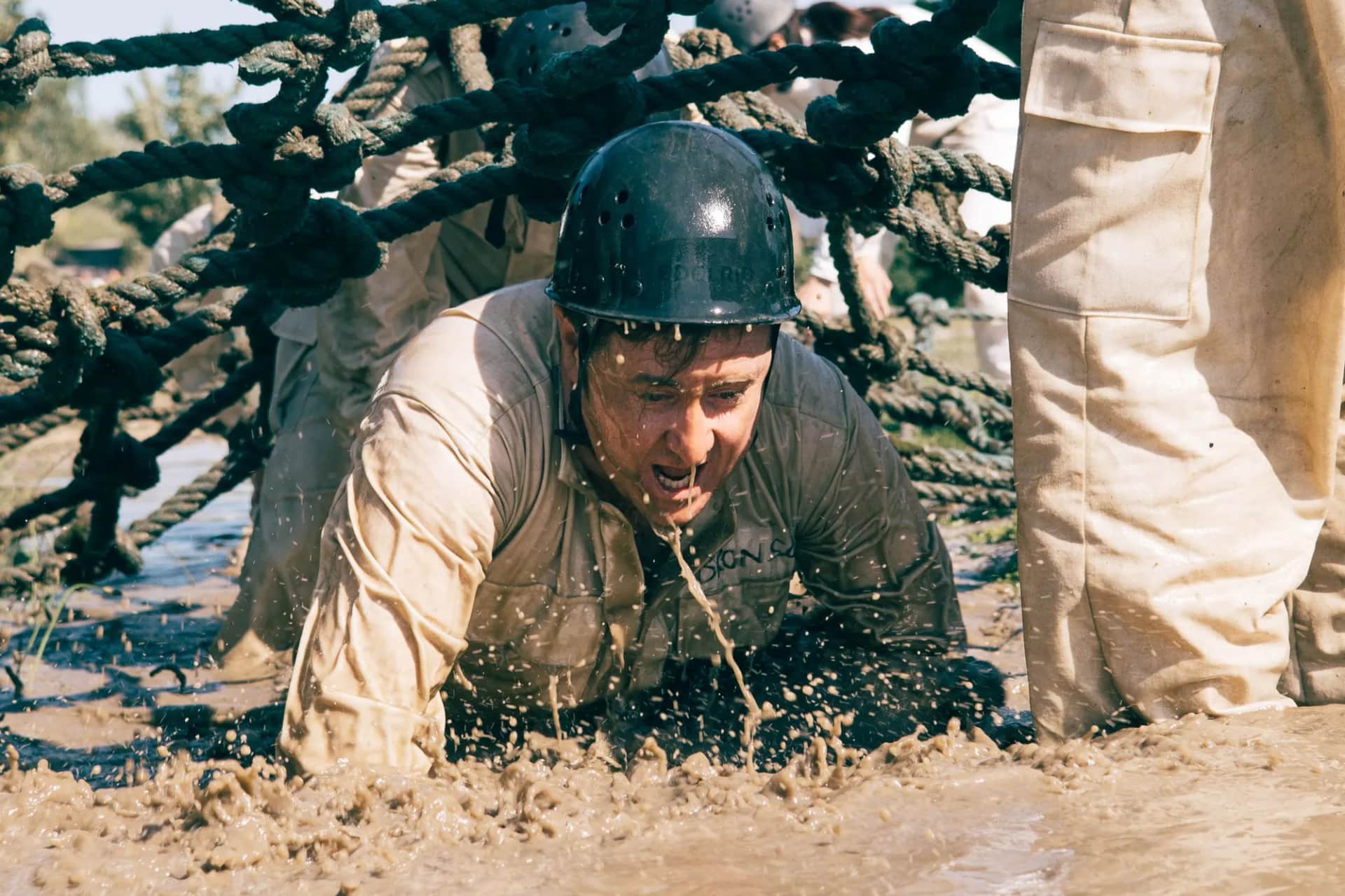 What conditions make experiential learning actually change behaviour?. A man in a helmet hauling himself under rope netting through deep mud