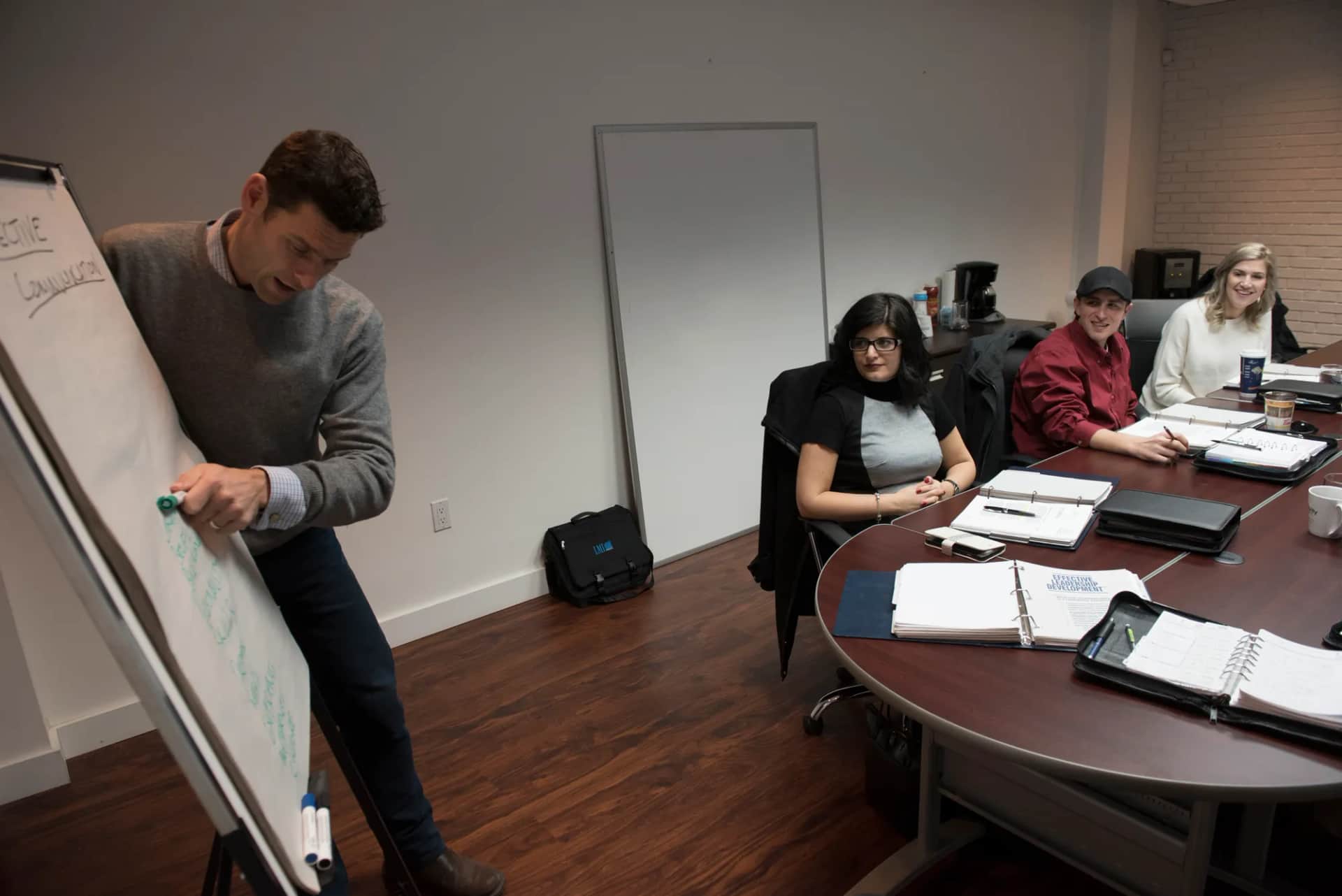 A facilitator writing on a flipchart while a small group watches from a boardroom table