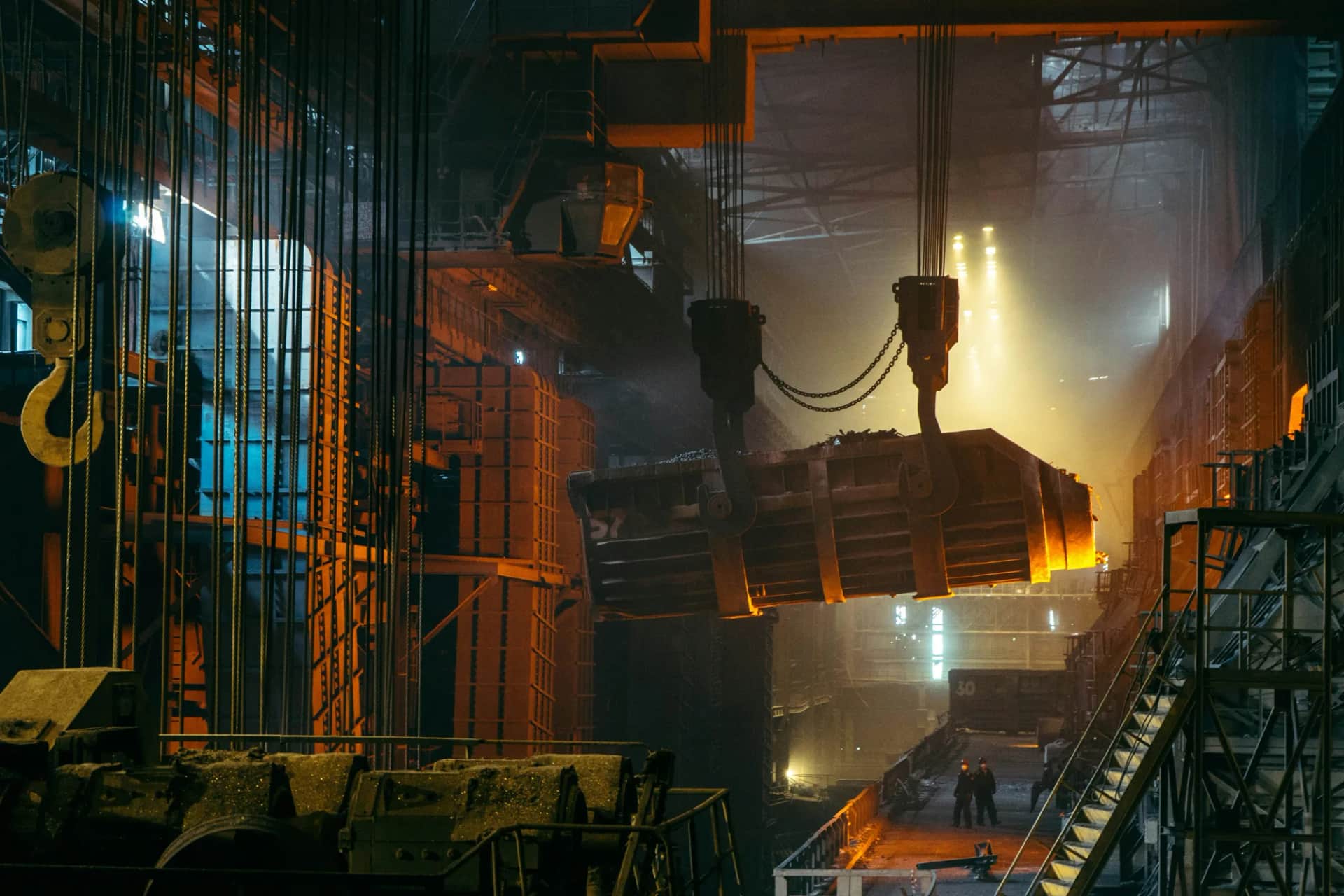 An overhead crane carrying a steel ladle through a foundry, two workers small on the floor below