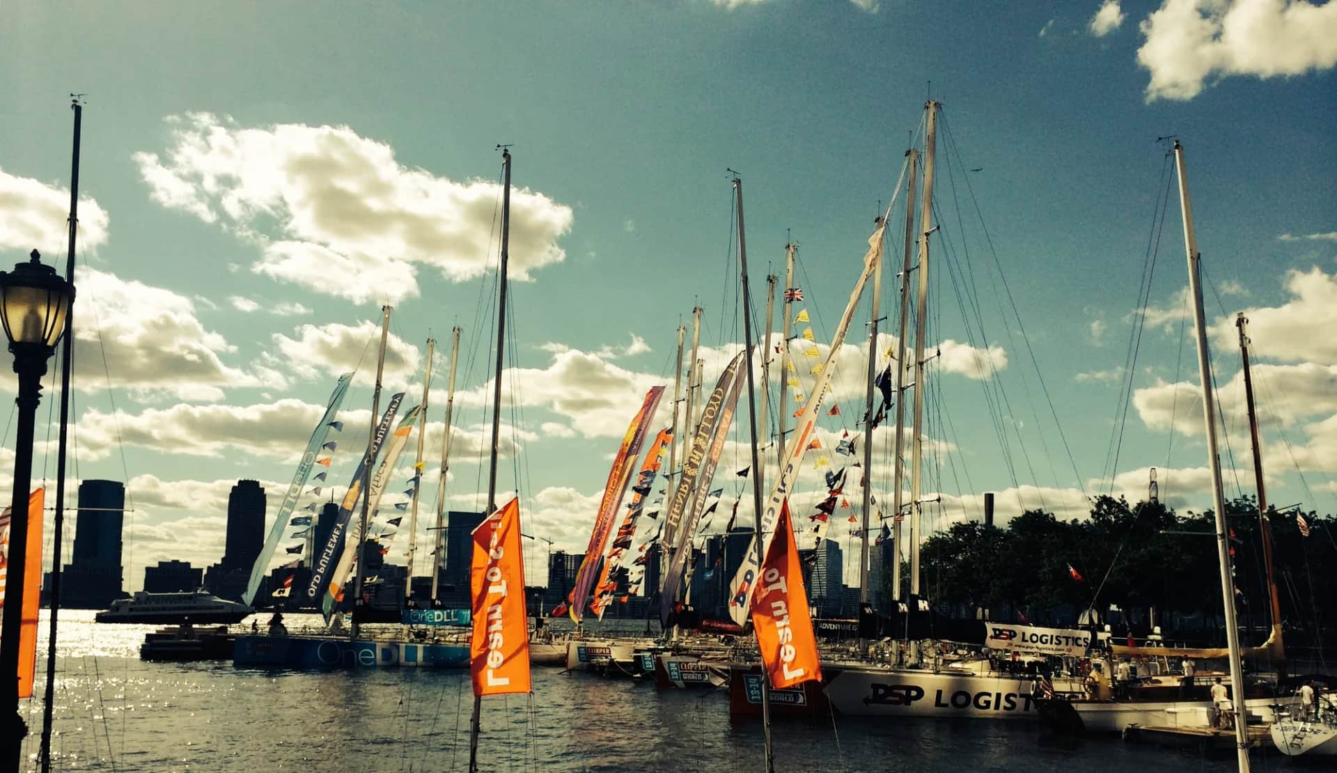 A line of ocean racing yachts moored together, pennants flying, a city skyline behind