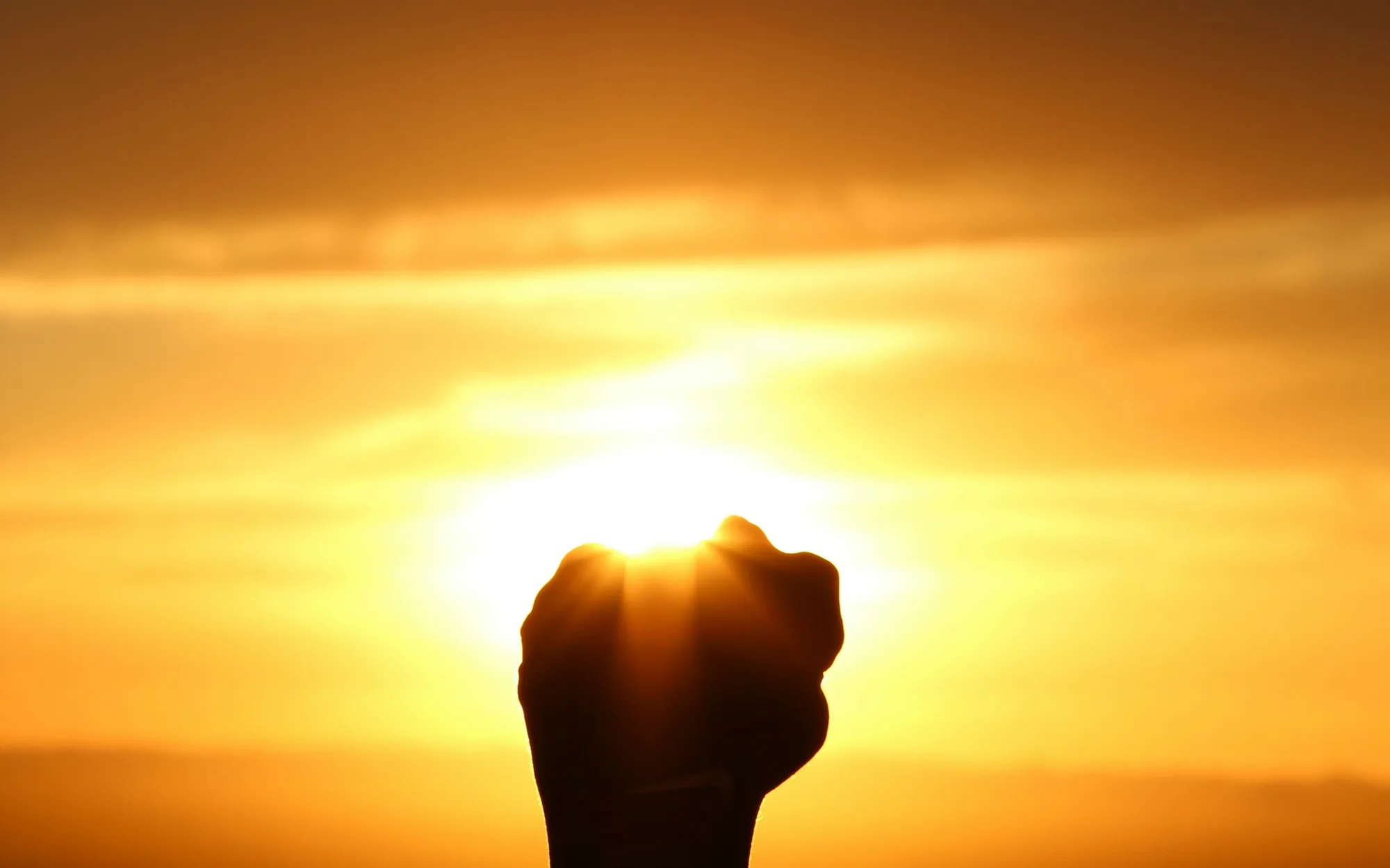 A raised fist in silhouette against a sunrise, the sun breaking through the fingers