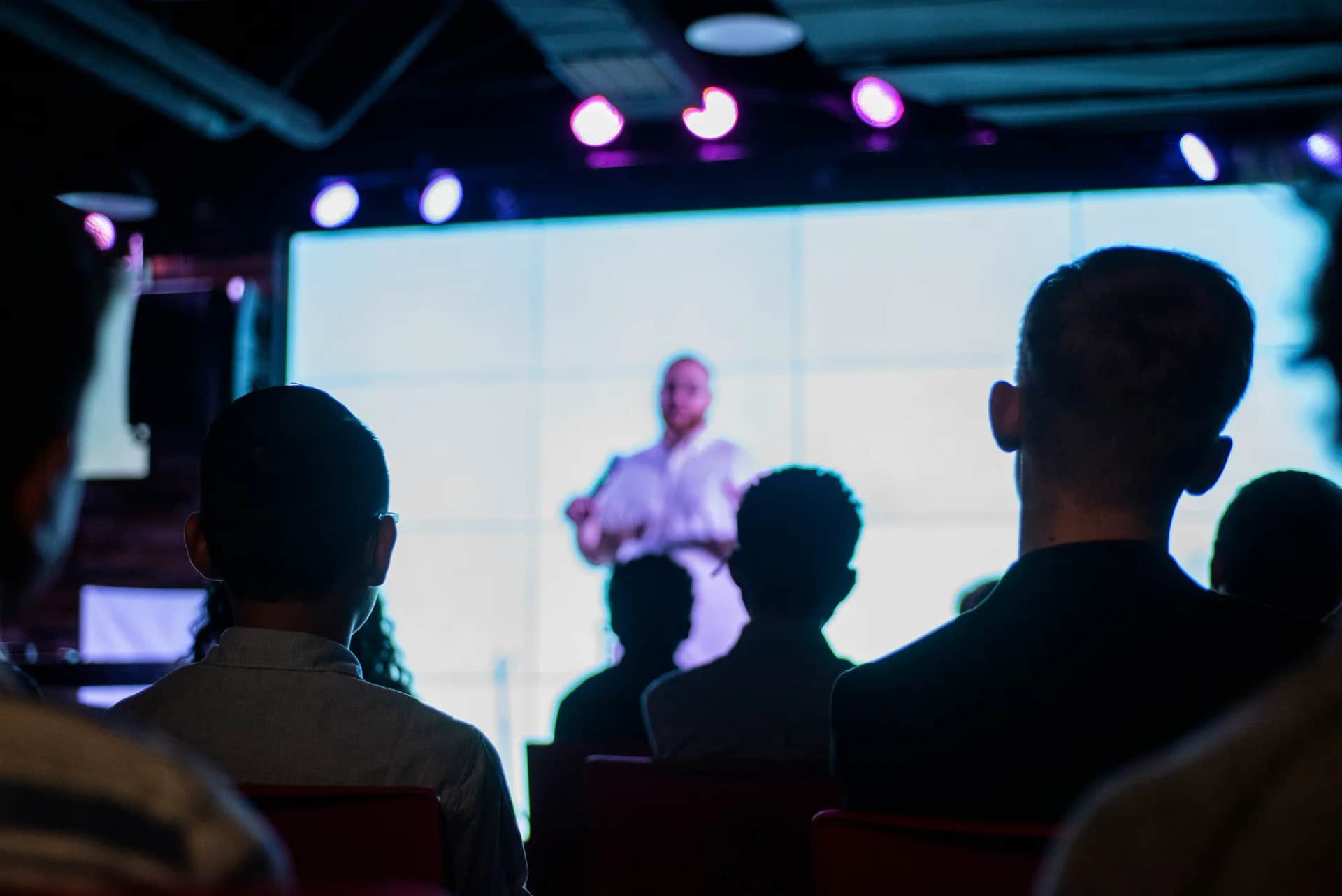 A presenter on a lit stage in front of a large screen, seen over the heads of a seated audience