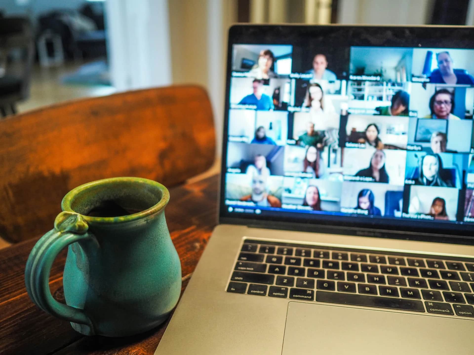 A laptop on a wooden table showing a video call full of participants, a mug of coffee beside it